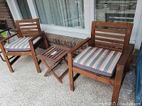 Two brown stained wooden outdoor armchairs with striped cushions facing forward, and a small foldable matching wooden stool placed between them on a patio surface in front of a window and brick wall.