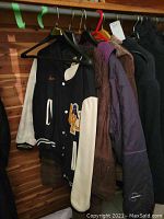 Black varsity style jacket with white leather sleeves hanging on rack among other coats. Shows general front view of jacket.