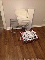 Full view of wire rack with stacked plastic storage bins on top, white boards behind, and red basket filled with box cutters on the floor.