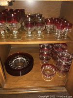 Shelved ruby flashed King's Crown pattern glasses and plates including stemmed glasses with and without gold trim and dessert bowls.
