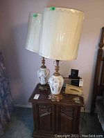 Two matching porcelain lamps with floral gold designs and brass bases placed on a wooden cabinet, shades wrapped in plastic.