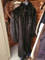Photo of the full length dark brown mink fur coat hanging on a hanger in a wooden closet, showing the coat's overall appearance and length.