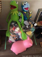 Photo of two large plush toys, Kermit the Frog (green) and Gonzo (blue), sitting on top of a clear plastic bag filled with various smaller plush toys; pink pig toy in front.