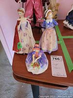Photo capturing three Royal Doulton lady porcelain figurines on a table with lot label 766 visible