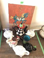 Photo showing six teapots, one cup, and coffee pot arranged on a surface with a painting of carousel horse behind them.