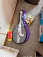 Photo of two tennis racket cases, black and purple, and a container and box of tennis balls.