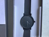 Close-up photo of the watch face, displaying the gray dial with minimal white markers and gray case with gray leather strap.