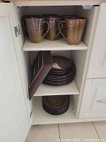 Photo of three large coffee mugs with brown/tan gradient and textured grid pattern on the top shelf of white cabinet.