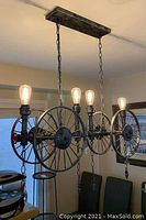 Front angled view of industrial chandelier showing three large spoked wheels horizontally mounted, four Edison bulbs lit, two metal chains attaching chandelier to ceiling plate.