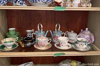Shelf view of 9+ teacups with saucers and 3 teapots with floral, green, blue, and white colorings