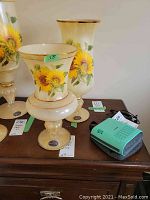 Two cream-colored Murano glass vases with hand-painted sunflowers and gold trim sitting on a wooden table with tags and a small green electronic device beside them.