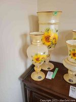 Two Murano glass vases with sunflower paintings and gold trim on a wooden surface.