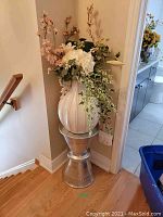 Full view of metal plant stand supporting a white ceramic vase filled with faux floral arrangement including white hydrangeas and trailing ivy.