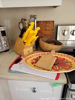 Overall view of countertop showing J.A. Henckels knife block with yellow handle knives, wooden salad bowl, decorative tray, cutting boards and utensils container