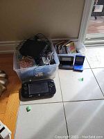Wide view of plastic bin containing cables, Wii console, Wii U GamePad on floor and two handheld devices