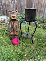 Front view of carved wood bear figure, small pink planter, and wrought iron plant stand in a garden area with grass and wooden fence background.