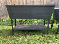 Full front and side view of the black resin elevated planter with metal legs and shelf underneath on grass against a fence.