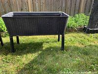 Full view of rectangular wicker style resin planter with metal legs placed outside on grass.