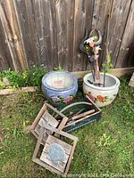 View of two large, round ceramic planters with floral decorations set on grass against a wooden fence, showing the garden plaques and garden tools in a basket in front.
