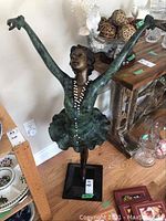 Full view of bronze-tone female dancer statue with arms raised, wearing pearl necklace, standing on black base, surrounded by household items.