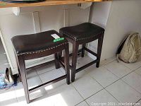 Pair of dark brown leather-top stools with studded detail and solid wood frames positioned under a kitchen island counter on a tiled floor.