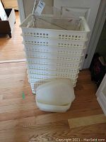 Photo showing a stack of about eight white plastic storage bins or hampers with square ventilation holes, a couple of clear plastic bins on top, and another white bin on the floor in front.