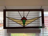 Front view of rectangular stained glass sun catcher with geometric red, green, and amber colored glass pieces in a wooden frame, hanging in front of a window.