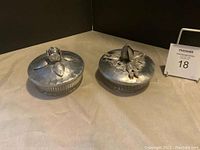 Two antique glass powder dishes with detailed hammered metal lids, placed side by side on a flat surface, shown from above.