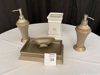 Photo of gold-tone soap pumps, soap dish, and white ceramic toilet brush holder on round white table.