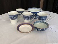 Set of four mugs in white with blue floral decorative patterns around the upper half and handles, placed together with one flat plate with orange and blue rim pattern and two bowls, one large and one small, both in blue and white with floral motifs.