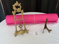 Two brass stands, one large ornate and one smaller, with a bright pink rolled yoga mat behind them.