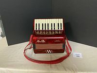 Front view of child-size red accordion with white piano-style keyboard and black buttons, candy apple red finish visible. Missing black buttons apparent.