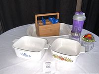 Photo showing two Corning Ware casserole dishes with different patterns, bamboo utensil holder with blue utensils, purple water bottle, jam jar with fruit lid, and the large white lasagna pan in back.