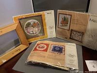 Photo of three cross stitch and crewel embroidery kits still in packaging next to a wooden stretcher bar.