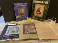 Five cross stitch and crewel kits displayed together. The kits are opened with visible charts and fabric. Visible brands include Dimensions and Mary Baxter St. Clair.