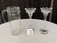 Front view of two crystal candle holders and a clear glass water pitcher on a round white table.