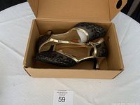 Pair of glitter black and gold dancing shoes in cardboard box on white tablecloth showing side and overhead views.