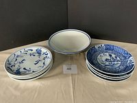 Photo of 4 oval white plates with blue floral and butterfly patterns, 4 small round plates with blue scenic designs, and 4 large bowls with blue trim and patterns arranged on beige cloth.