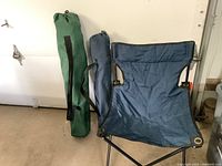 Three folding camping chairs, two blue and one green, with their respective carrying bags placed against a white wall.