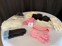 Winter accessories on white round table showing hats, scarves, gloves, and leg warmers in predominantly white, pink, black colors.