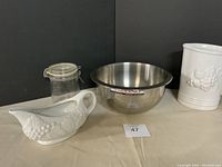 Full view of four kitchen items on beige cloth background: stainless steel mixing bowl labeled Paderno, white ceramic milk pitcher, white ceramic gravy boat, clear glass jar with latch lid.