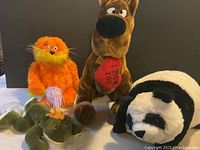 Photo showing four stuffed toys including Scooby-Doo, a panda, an orange furry creature, and a green dragon