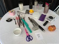 Photo showing a variety of kitchen items including bottles, tongs, citrus juicer, and other utensils arranged on table