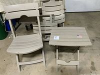 Two beige plastic folding patio chairs and two matching folding tables stacked leaning against a wall, showing the slatted design and flat folding capability.