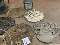 Photo showing five decorative patio stones with various designs including welcoming message, flowers, birds, and celestial motifs, alongside a watering can and metal jug.