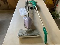 Shark steam mop and green Swiffer Sweeper Vacuum cleaner displayed on a table alongside an extra mop pad