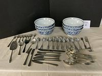 Photo showing 6 white ceramic soup bowls with blue floral pattern and multiple Rogers Community silverplate flatware pieces arranged on white cloth background.