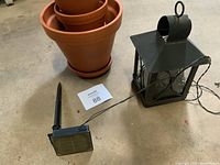 Four stacked terra cotta pots with saucers next to a black metal lantern with solar panel and attached fairy lights, placed on concrete floor.
