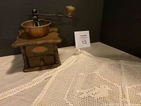 Photo of vintage wood coffee grinder with metal handle crank and drawer alongside hand made crochet apron.