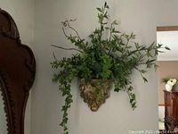 Front view of artificial green leafy plant arrangement in gold/brass decorative wall bracket mounted on wall.